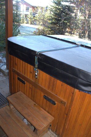 Un bain à remous en bois, avec un banc devant, installé à l’extérieur, entouré d’arbres et avec un bâtiment en arrière-plan.