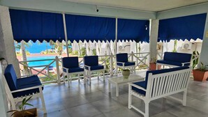 Un espacio de estar al aire libre cubierto, con mobiliario blanco y cojines azules, con vistas a una piscina y zonas verdes.