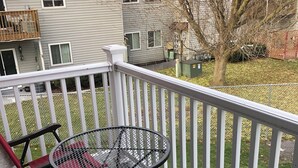 Um terraço com mesa e cadeiras de metal, rodeado por um corrimão branco, com vista para um jardim gramado com uma árvore e um prédio ao fundo.