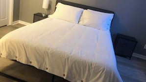 Uma cama bem arrumada com roupa de cama branca e cabeceira escura em um quarto com parede lisa e uma pequena mesa de cabeceira com abajur.