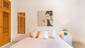 A bedroom with a bed, pillows, a nightstand with a lamp, a wall-mounted painting, and wooden shutters.