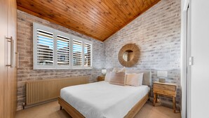 A bedroom with a wooden ceiling, brick walls, a large bed, a bedside table, and a round mirror.
