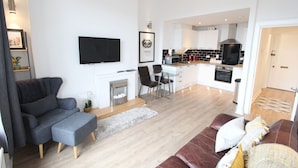A modern living room with a grey sofa, a brown leather couch, and a dining area with a glass table and chairs. A kitchen with a black stove and white cabinets is visible in the background.