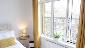 A hotel room with a bed, a bedside table with a lamp, a window with curtains, and a view of buildings outside.