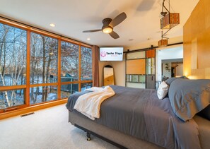 A modern bedroom with a large window, a bed, a ceiling fan, and a TV displaying 'Sacha Stays of Wisconsin'.