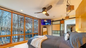 A modern bedroom with a large window, ceiling fan, and a flat-screen TV. - Luxe Lakefront Estate near Minneapolis, Hot Tub, Sauna, Kayaks, Sleeps up to 20 (Baldwin)