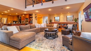 A spacious living area with a large sectional sofa, a round coffee table, and a fireplace. A kitchen is visible in the background, and there is a balcony with a railing. - Luxe Lakefront Estate near Minneapolis, Hot Tub, Sauna, Kayaks, Sleeps up to 20 (Baldwin)