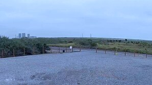 A gravel parking area with a fence, a small structure, and a distant view of buildings.