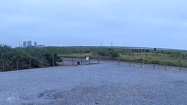 Un espace de stationnement recouvert de gravier, délimité par une clôture, avec une petite construction et une vue lointaine sur des bâtiments.