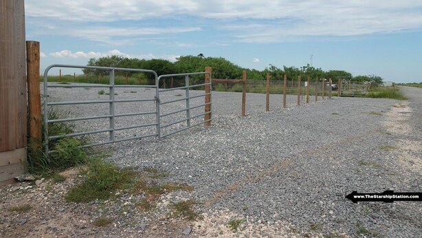 A gravel path leading to a metal gate, with a wooden structure on the left and a fence in the background.