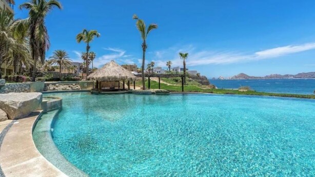 Una piscina azul cristalina con una estructura de techo de paja, palmeras y vista al mar.