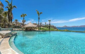 Ein klares, blaues Schwimmbecken mit einer strohgedeckten Struktur, Palmen und Blick auf das Meer.