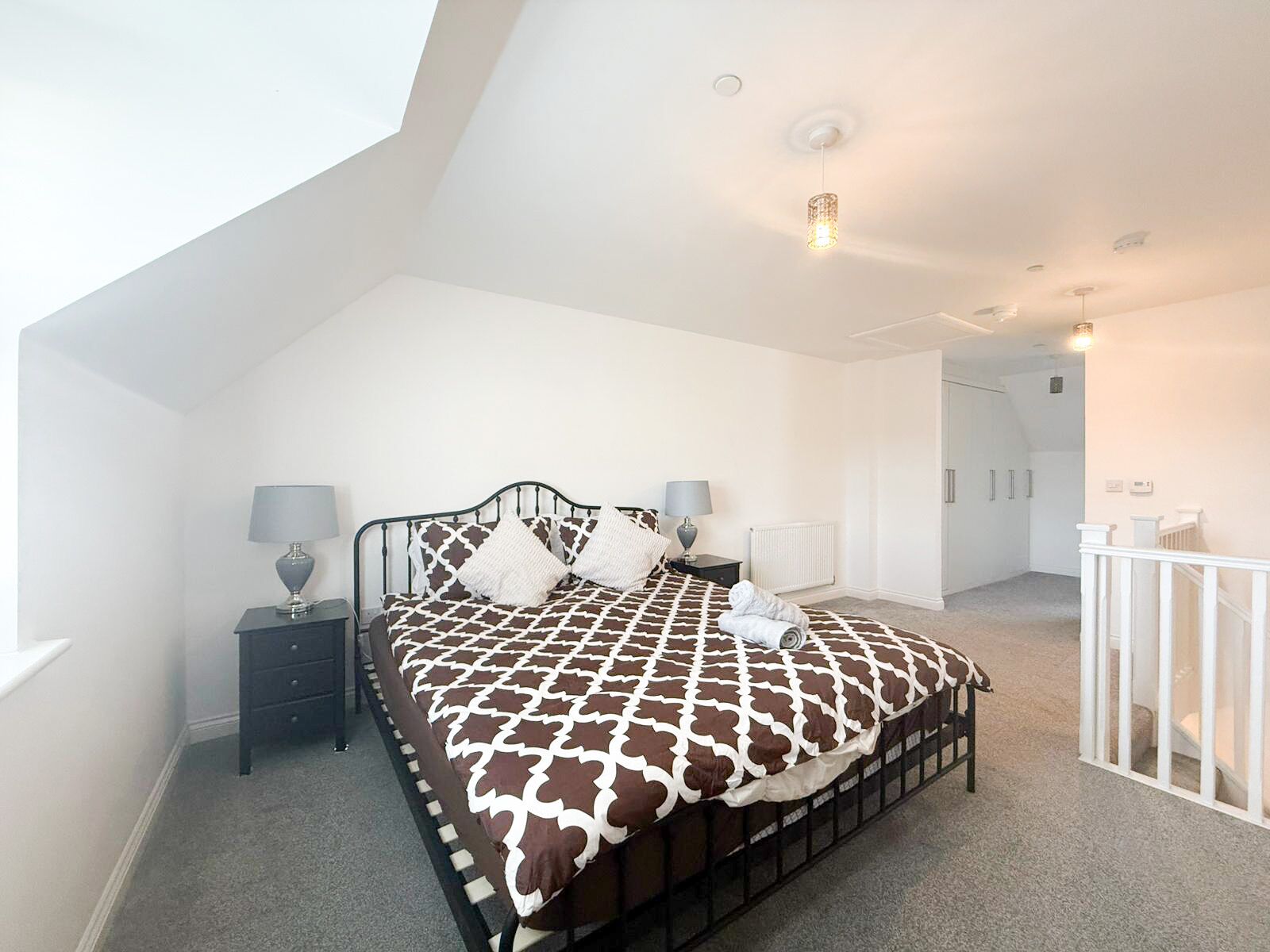 A bedroom with a high ceiling, a bed with a patterned bedspread, two bedside tables with lamps, a radiator, and a white railing.