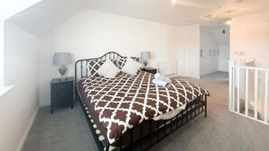 A bedroom with a high ceiling, a bed with a patterned bedspread, two bedside tables with lamps, a radiator, and a white railing. - Your Bromley Experience  (Bromley)