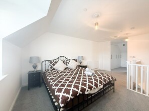 A bedroom with a high ceiling, a bed with a patterned bedspread, two bedside tables with lamps, a radiator, and a white railing. - Your Bromley Experience  (Bromley)