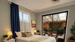 Una camera da letto con un letto grande, un comodino, una lampada e vista su un patio esterno con sedie e piante.