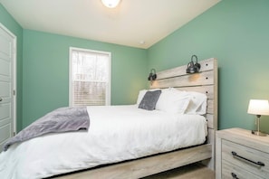 A bedroom with a bed, a nightstand, a lamp, a window with blinds, and a door.