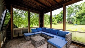 A covered outdoor seating area with a sectional sofa and ottomans, large windows, and a wooden ceiling. - New Remodel on Black River with Hot Tub, Kayaks, Gym, Fireplace, & Game Room (South Haven)