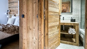 Ett modernt sovrum med en trägarderob, en säng med ett brunt överkast, en sänglampa och ett badrum med ett träskommod och en spegel.