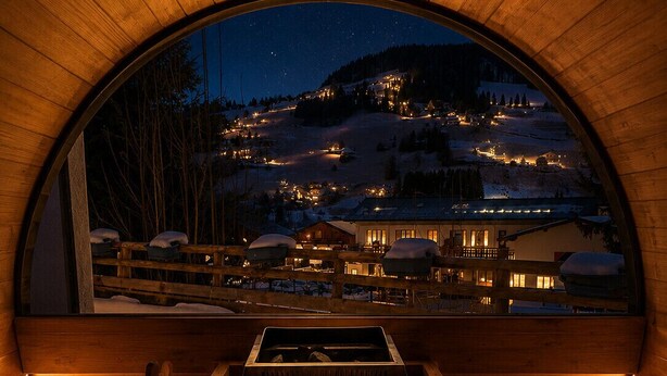 Eine Holzsauna mit Blick auf ein schneebedecktes Dorf bei Nacht.