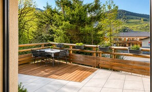 Une terrasse en bois, agrémentée d’une table et de chaises, entourée d’une balustrade en bois, et offrant une vue panoramique sur un paysage verdoyant, avec des arbres et un bâtiment visible au loin.