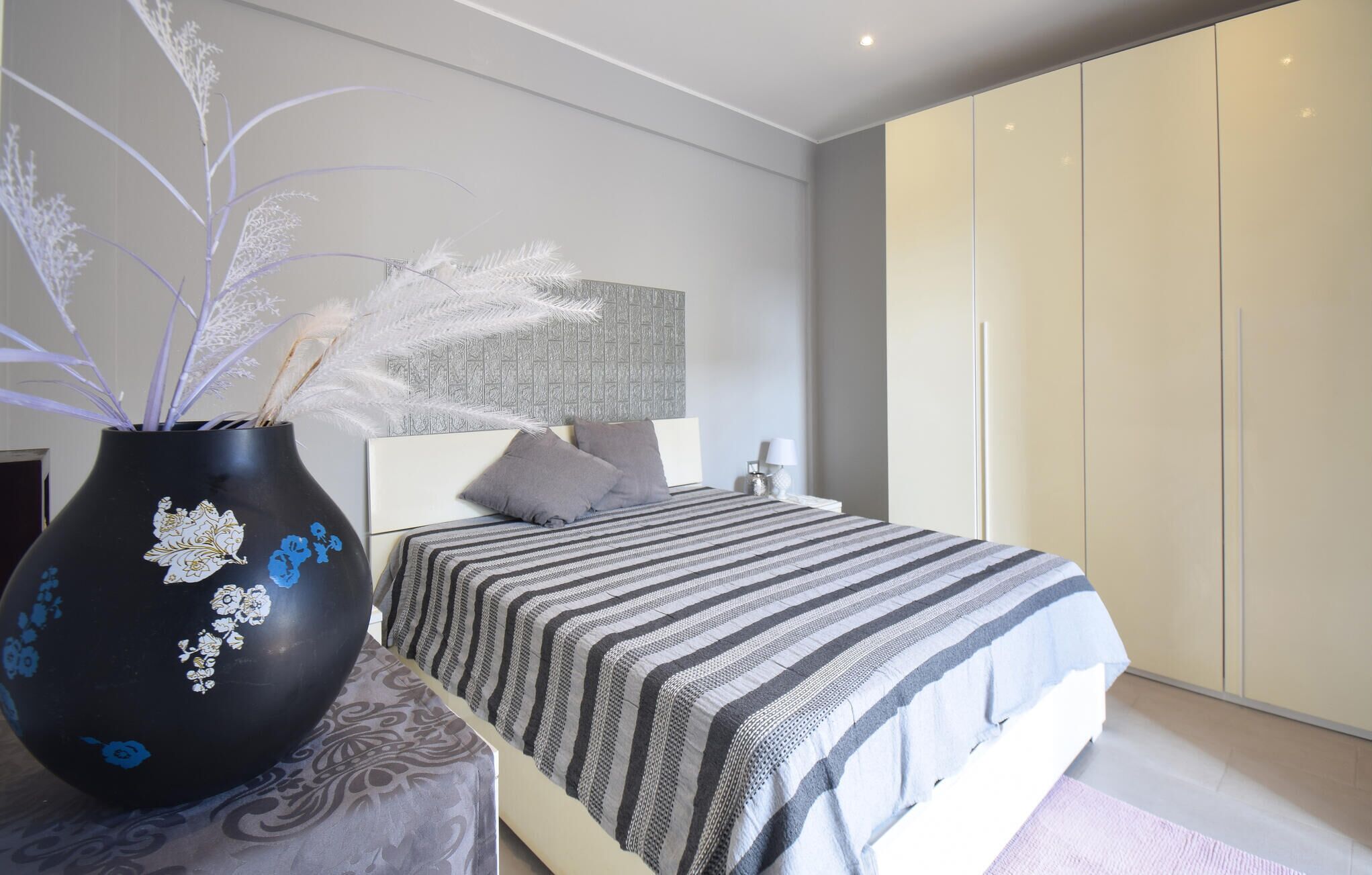 A modern bedroom with a bed, a nightstand, a large wardrobe, and a decorative vase with dried plants.