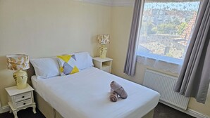 A hotel room with a bed, two bedside tables, a lamp, a window with curtains, and a view of buildings outside. - Large 6-Bed House | 2 Bathrooms | Parking | Garden (Bristol)