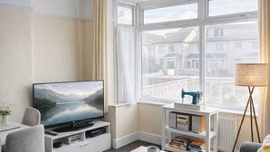 A modern living room with a television, white furniture, a coffee table, a sofa, a lamp, and a view of buildings through large windows. - Large 6-Bed House | 2 Bathrooms | Parking | Garden (Bristol)