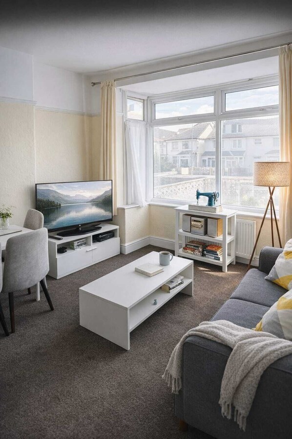 Un salon moderne doté d’un téléviseur, de meubles blancs, d’une table basse, d’un canapé, d’une lampe et offrant une vue sur les bâtiments à travers de grandes fenêtres.