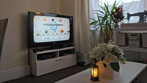 A television displaying the Mario Kart Wii game menu, a white entertainment unit with a sewing machine, and a vase with white flowers on a table. - Large 6-Bed House | 2 Bathrooms | Parking | Garden (Bristol)