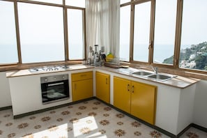 Uma cozinha com armários amarelos, fogão e pia. Possui grandes janelas com vista para o mar e uma encosta.