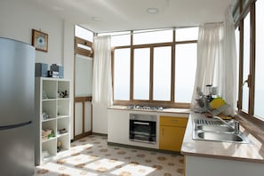 Uma cozinha compacta com fogão a gás, forno e pia. Possui uma janela com cortinas, geladeira e uma prateleira com utensílios de cozinha.