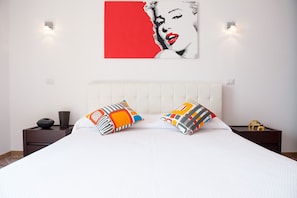Quarto com cama branca, duas almofadas coloridas, cabeceira branca e obra de arte vermelha e branca na parede.