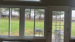 A room with large windows overlooking a grassy area with a wooden fence and trees. - Charming 3-bedroom detached cottage on horse farm near Lexington and Georgetown. (Paris)