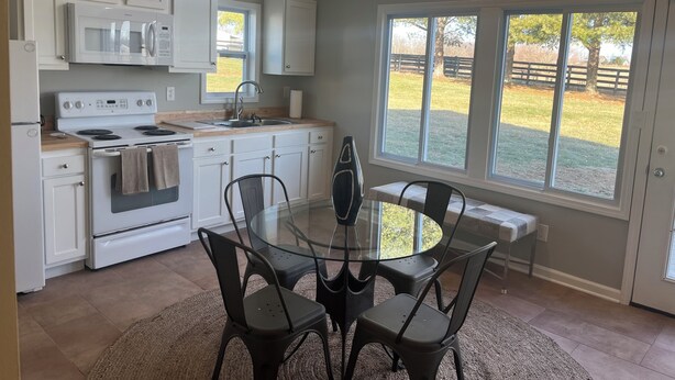 A modern kitchen with white cabinets, a stainless steel refrigerator, a stove, and a sink. There is a glass dining table with four chairs. Large windows provide a view of a grassy yard. - Charming 3-bedroom detached cottage on horse farm near Lexington and Georgetown. (Paris)