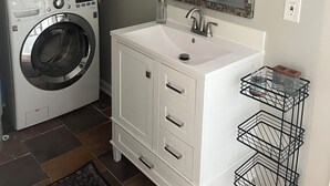 A laundry area with a front-loading washer and dryer, a white vanity with a mirror, and a black metal shelf unit. - Charming 3-bedroom detached cottage on horse farm near Lexington and Georgetown. (Paris)
