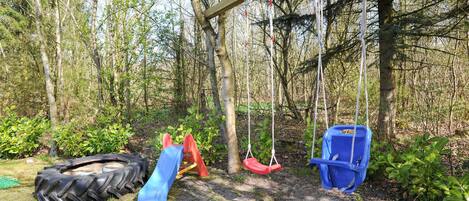 Une aire de jeux avec une balançoire faite d’un pneu, un toboggan et un ensemble de balançoires, le tout situé dans une zone boisée.