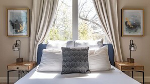 Un dormitorio con una cama grande, dos mesitas de noche y una ventana con cortinas. Se observan dos cuadros enmarcados en las paredes.