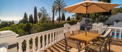 Una terraza con una mesa y sillas, una sombrilla color beige, y vistas al mar y a los árboles.