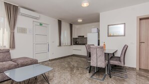 Een moderne woonkamer met een bank, een tafel met glazen blad en een eethoek met stoelen en een tafel. In de achtergrond is een keuken met kastjes en een koelkast te zien.