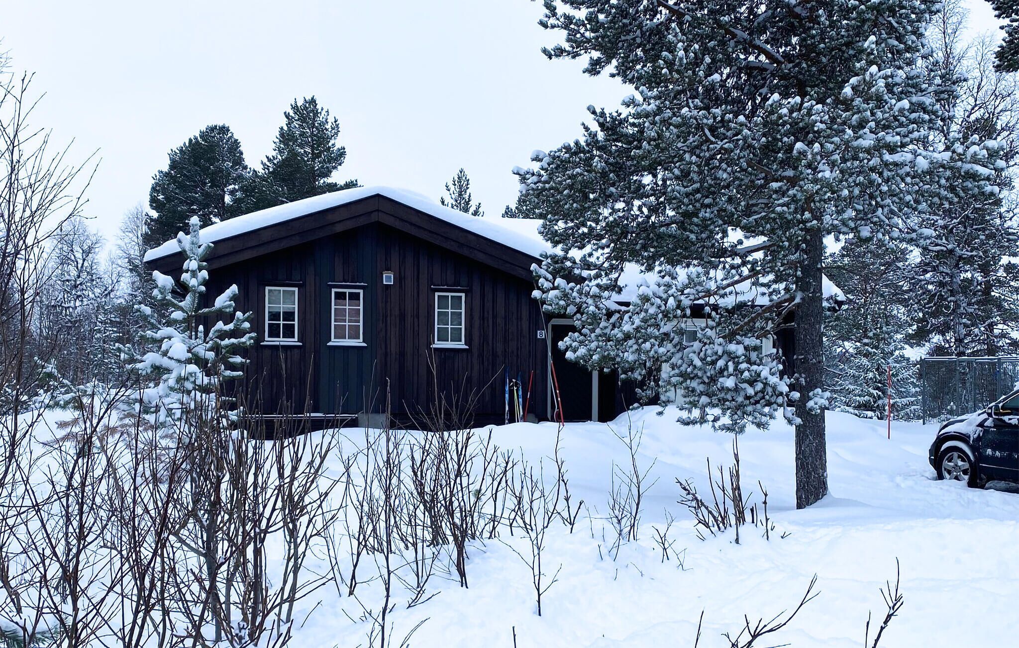 A dark wooden cabin with a snow-covered roof, surrounded by snow-laden trees and a car parked nearby.