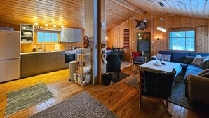 A cozy cabin interior with wooden walls and floors, a kitchen with modern appliances, a dining area, and a living room with a large window.