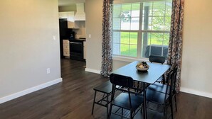 A dining area with a table and chairs, a window with curtains, and a view of a kitchen and outdoor area.