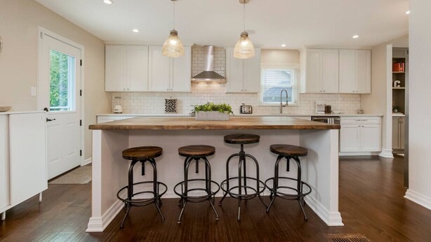 A modern kitchen with white cabinets, a wooden island with stools, pendant lights, and a large window. - Kid-Friendly 4 Bedroom Home w/ Heated Private Pool (Overland Park)