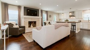 A modern living room with a fireplace, a sofa, a TV, and a kitchen area with white cabinets and a bar stool. - Kid-Friendly 4 Bedroom Home w/ Heated Private Pool (Overland Park)