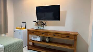 Quarto com escrivaninha de madeira, televisão de tela plana na parede, pequeno frigorífico e cama com colcha verde.