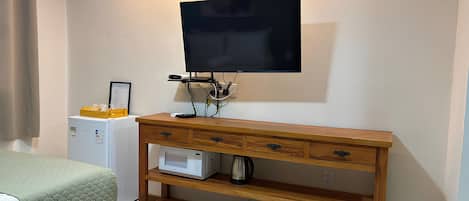 Una habitación con un escritorio de madera, un televisor de pantalla plana en la pared, un refrigerador pequeño y una cama con colcha verde.