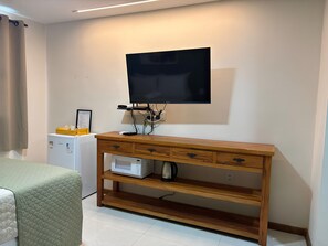A room with a wooden desk, a flat-screen TV mounted on the wall, a small refrigerator, and a bed with a green quilt. - Suítes Brisa do Mar (Búzios)