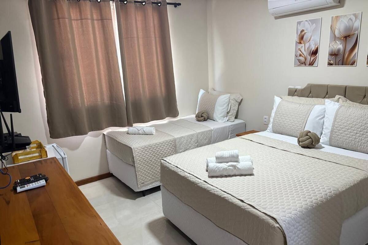 A hotel room with two single beds, a larger bed, a wooden desk with a remote control, and a television.