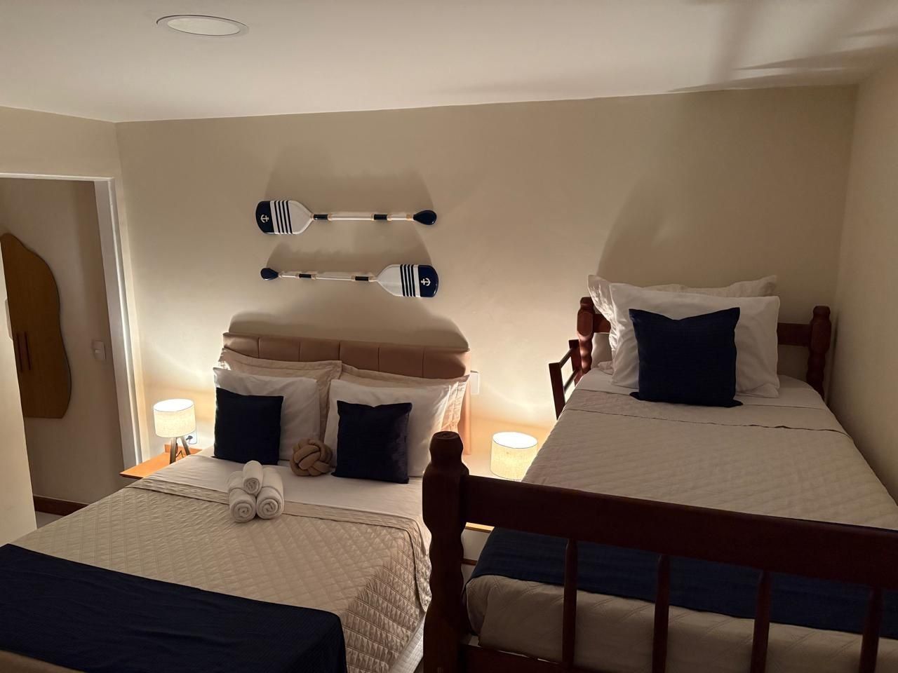 Habitación de hotel con dos camas, cada una con cabecero de madera, una mesita de noche con lámpara y un perchero con toallas.